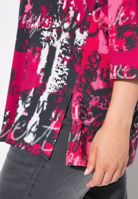 Kleurvolle tuniek met een roze, zwarte en witte grafische motif, korte mouwen en zijsplitten, gemaakt van een gestructureerde stof.