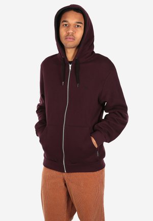 Junge Person trägt einen dunkel kastanienbraunen Hoodie mit Reißverschluss, die Hände in den Taschen, und hellbraune Hosen, steht vor einem einfarbigen Hintergrund.