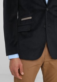 Marinblå texturerad kavaj med två knappstängning, sidoficka med ljusa accentsträck, kombinerad med en ljusblå skjorta och beige byxor.