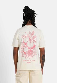 Crème katoenen t-shirt met een roze fruitafbeelding van sinaasappels en bladeren, met de tekst "GENIET VAN DE SMAAK" eronder. Korte mouwen, ontspannen pasvorm.