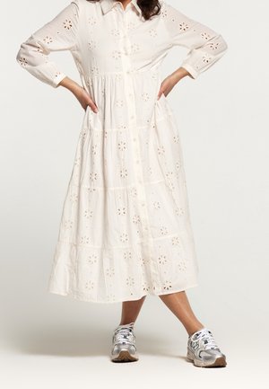 Femme en robe midi blanche boutonnée avec découpes florales et baskets argentées, debout les mains sur les hanches sur fond blanc.