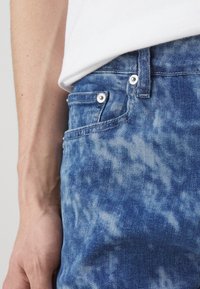 Falmet blå denimjeans med et subtilt sky-lignende mønster, som har sølvfarget detaljer og en frontlomme. Nærbilde med en hånd som hviler i nærheten.