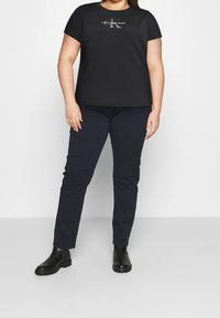 Camiseta de algodón negra con cuello redondo y mangas cortas, que presenta un logo plateado. Combinada con pantalones ajustados azul oscuro y botas negras hasta el tobillo.