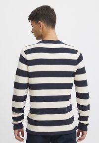 Pulover lung cu mânecă lungă în dungi bleumarin și crem, textură tricotată, guler rotund, detaliu cu etichetă pe lateral, croială relaxată. Vedere din spate care arată dungi orizontale.