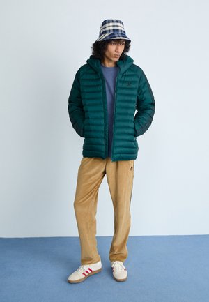 Teal doorgestikt jack met een capuchon, gedragen over een blauw shirt. Beige corduroy broek en witte sneakers met rode strepen. Blauwe geblokte bucket hat.