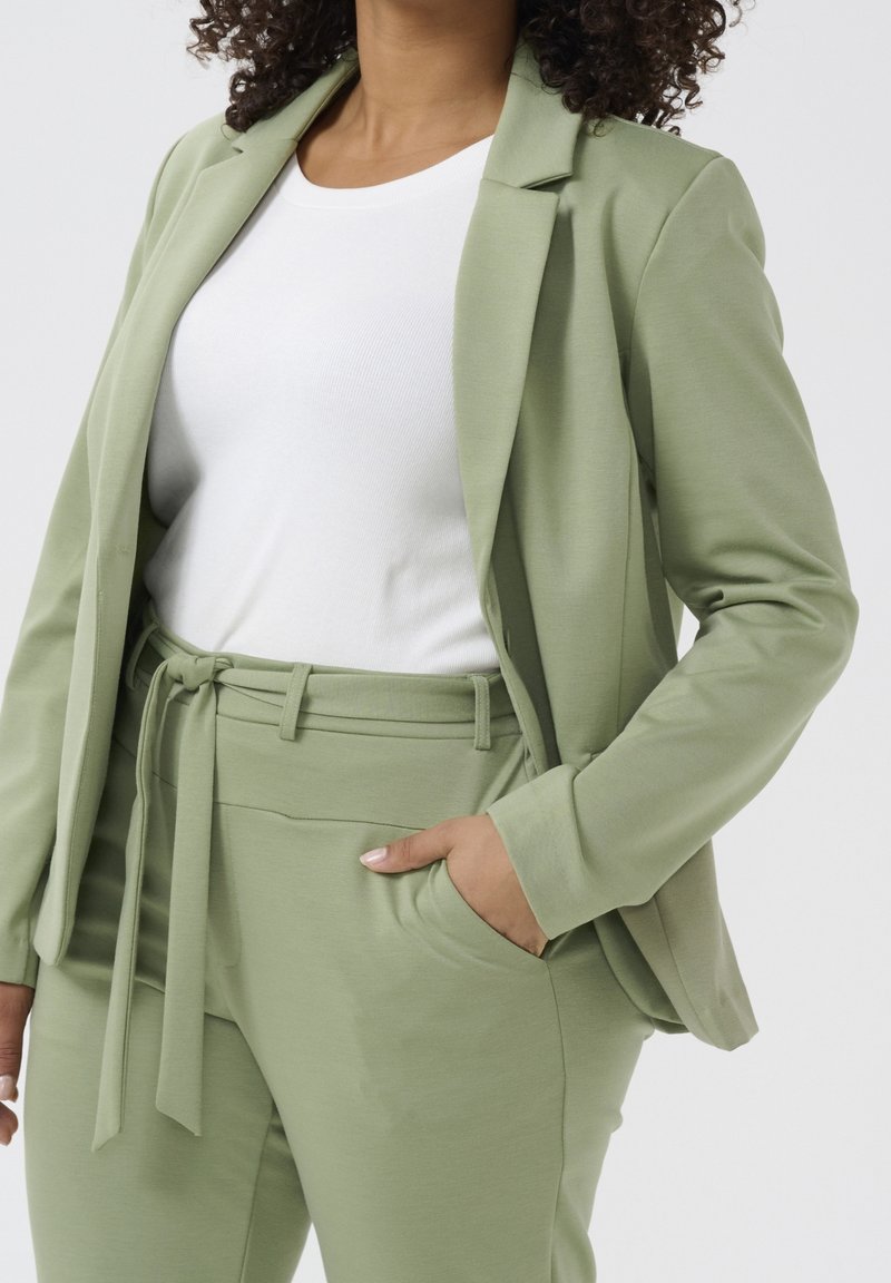 Femme en tailleur vert clair cintré avec ceinture à nouer portant un top blanc moulant, main dans la poche sur fond uni.
