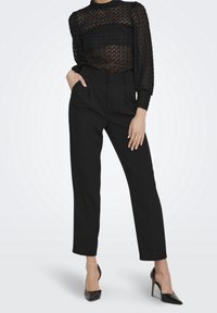 Blusa in pizzo nero con un motivo testurizzato, maniche lunghe e collo alto, indossata sopra pantaloni neri sartoriali e scarpe elegante con tacco alto nere.