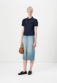 Marineblå skjorte med knapper og puffede ærmer, lysblå denim blyantskirt med forslidser, sorte sko og en lille vævet brun håndtaske.