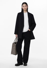 Svart oversized blazer over en hvit strukturert topp, kombinert med svarte bukser og kraftige svarte støvler. Holder en svart og beige rutete veske.