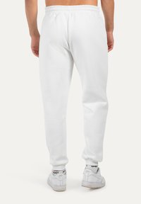 Bílé tepláky s elastickým pasem, manžetami v oblasti kotníků a hladkým povrchem. Nošeny s bílými teniskami s černými akcenty.