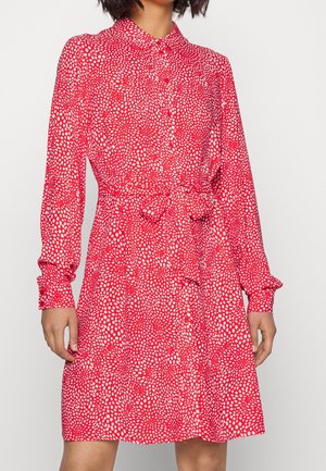 Woman wearing a red long-sleeve dress with white irregular dots, buttoned front, collar, and tied waist belt, standing against a white background.