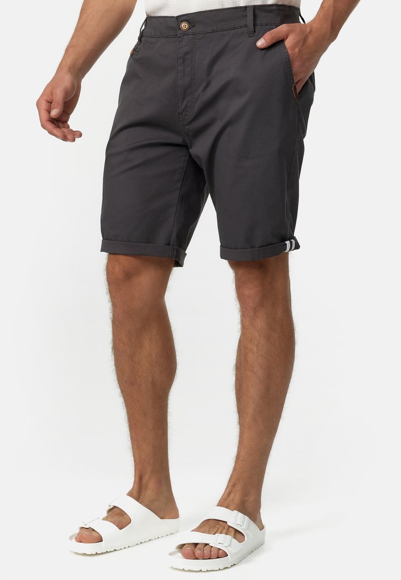 Homme portant un short gris foncé retroussé et des sandales blanches à double bride, debout avec une main dans la poche.