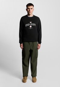 Felpa nera con logo "LYLE & SCOTT", pantaloni cargo verde oliva e scarpe slip-on color cammello. Tessuto liscio, vestibilità rilassata e design minimal.