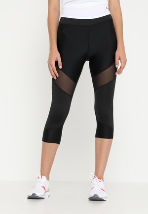 Frau trägt schwarze gekürzte Leggings mit transparenten diagonalen Einsätzen und weißen Sportschuhen, steht vor einem schlichten weißen Hintergrund.