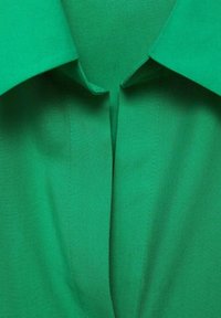 Vue rapprochée d’un col de chemise en tissu vert avec des coutures propres et une petite ouverture en forme de V à l’encolure.