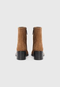 Stivaletti in suede marrone alla caviglia con punta arrotondata e tacco in legno nero, dotati di chiusura con cerniera sul retro per un facile indosso.