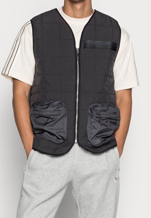 Schwarze gesteppte Weste mit Reißverschluss, gepolsterten Taschen und strukturierten Schulterakzenten. Kombiniert mit einem hellen, gestreiften T-Shirt.