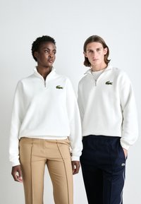 Baltos zip-up džemperiai, pagaminti iš minkštos medžiagos, su žaliu Lacoste logotipu ant krūtinės; derinami su BEIGE ir tamsiai mėlynomis kelnėmis.