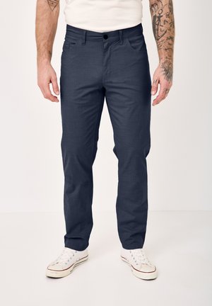 Man wearing navy blue straight-leg pants and white sneakers, standing against a plain white background, tattoos visible on forearms.