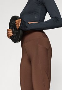 Zwarte top met lange mouwen en duimgaten, bruine legging met hoge taille voorzien van een geribbelde textuur en een zijzak, en een zwarte gewichtsschijf in de hand gehouden.