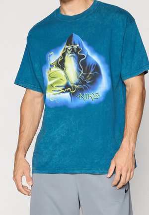 Homme portant un t-shirt bleu avec un graphisme d’un sorcier barbu tenant un ballon de basket lumineux avec des effets électriques et le logo Nike en dessous.
