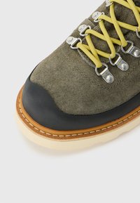Close-up of a hiking boot with olive green suede, yellow laces, metal eyelets, black rubber toe cap, and beige sole with stitching.