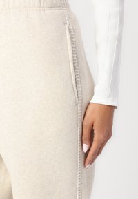 Beige Sweatpants mit einer weichen Textur, ausgestattet mit seitlichen Taschen und kontrastierenden Nähten entlang der Naht. Ein weißes, geripptes Langarmoberteil ist teilweise sichtbar.