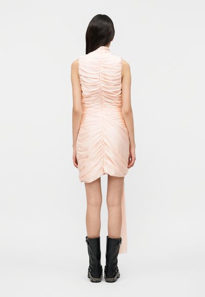 Γυναίκα με μακριά σκούρα μαλλιά, ντυμένη με αμάνικο ρόζ φόρεμα με σούρες και μαύρα μποτάκια μέχρι τη μέση της γάμπας, στέκεται γυρισμένη πλάτη σε απλό φόντο.