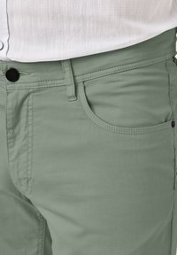 Hellgrüne Hose aus glattem Material, klassischem Five-Pocket-Design und schwarzem Knopfverschluss in der Taille. Sichtbare Nähte.