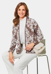 Cardigan à motifs marron et blanc avec une ouverture à l'avant, porté avec un top blanc et un pantalon clair. Tenant un sac à main couleur crème.