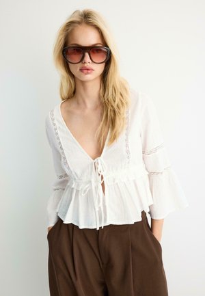 Blondynka w dużych brązowych okularach przeciwsłonecznych, biała bluzka z długim rękawem i wiązaniami oraz brązowe spodnie z wysokim stanem, stojąca na białym tle.