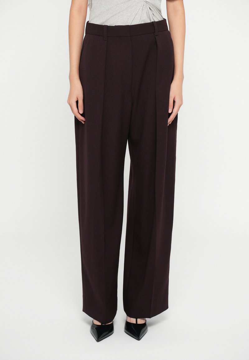 Pantaloni larghi color bordeaux con una vestibilità sartoriale, frontale pieghettato e passanti per cintura. Indossati con tacchi neri a punta. Tessuto liscio e strutturato.