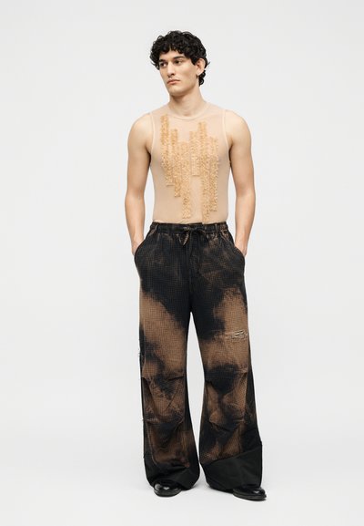 Άνδρας όρθιος με τα χέρια στις τσέπες, φορώντας αμάνικη μπεζ μπλούζα με υφασμένους κάθετους μοτίβους και φαρδιά μαύρα και καφέ παντελόνια με σχέδια.