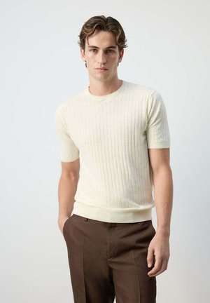 Giovane uomo con capelli corti e mossi che indossa una maglia a maglia color crema a maniche corte e pantaloni marroni su uno sfondo chiaro uniforme.