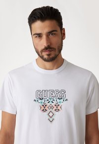Weißes Baumwoll-T-Shirt mit "GUESS" in schwarzer Schrift und einem geometrischen, in Türkis, Pfirsich und Schwarz gestickten Muster auf der Brust.