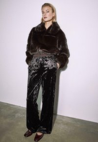 Žena v tmavohnedej umelej kožušine, čiernych zamatových širokých nohaviciach a tmavohnedých topánkach, stojaca pred jednoduchou svetlou stenou.