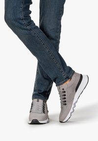 Scarpe sportive grigie con una tomaia testurizzata, suola bianca e nera, e occhielli prominenti per i lacci, abbinate a jeans blu scuro.