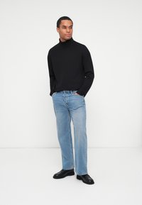 Maglione a collo alto nero, jeans dritti azzurri chiari e scarpe nere. Design semplice con una texture morbida, in piedi su uno sfondo bianco.