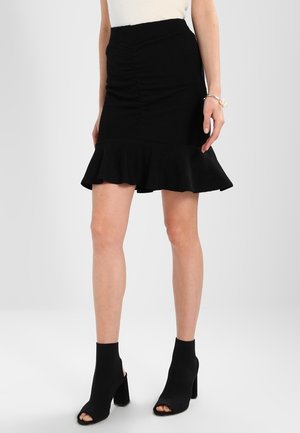 Femme portant une jupe noire froncée avec un ourlet à volants et des bottines noires ouvertes à talons, debout devant un fond clair uni.