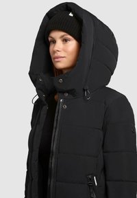 Vrouw in een zwarte gewatteerde jas met een grote capuchon en een zwarte gebreide beanie, die naar links kijkt tegen een effen achtergrond.