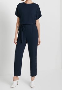Mörkblå jumpsuit med korta ärmar, cinchad midja och knytdetalj. Har en rak bendesign och slät tygstruktur. Matchas med vita klackar.