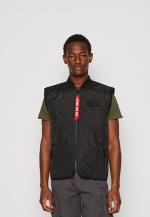 Zwarte gewatteerde vest met een hoge kraag, ritssluiting aan de voorzijde, zijzakken en een rode tag. Gemaakt over een groen shirt en grijze broek. Effen achtergrond.