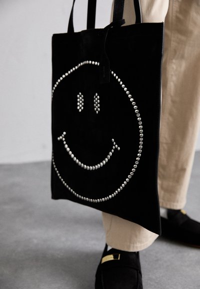 Geantă de umăr neagră cu un design de față zâmbitoare realizat din cristale argintii. Dispune de două mânere robuste și un material texturat, neted.