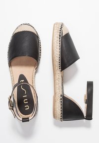 Espadrille de cuero negro con punta redonda, suela de esparto y correa para el tobillo. Confeccionada con un material texturizado en la parte superior y forro interior beige.