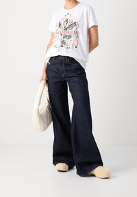 Bílý tričko s barevným grafickým designem; tmavě modré široké džíny; smetanová taška přes rameno; hnědé chlupaté obuv.