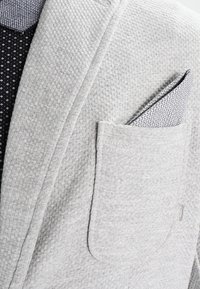 Grauer Blazer mit Struktur und Brusttasche, die ein gemustertes Einstecktuch zeigt. Darunter ein schwarzes Hemd mit Polka-Dot-Design.