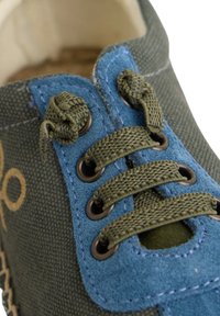 Zapatilla de lona con un cuerpo verde y acentos azules, ojales de metal y cordones de color verde oliva, con una superficie texturizada.