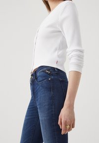 Cárdigan blanco de canalé con escote en V y cierre de botones, combinado con unos vaqueros oscuros de corte slim con una etiqueta de diseñador en el bolsillo trasero.
