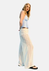 Un haut sans manches bleu clair avec un ourlet évasé et un pantalon palazzo blanc en dentelle présentant des motifs complexes et une coupe ample. Des tongs noires.