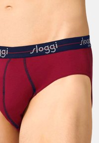 Dunkelrote Unterhose aus weichem Stoff mit einem marineblauen Bund, mit grauem "sloggi"-Logo und genähten Akzenten für ein strukturiertes Design.